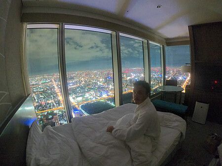 Persona seduta su un letto in una stanza d'angolo dell'Osaka Marriott Miyako Hotel con vista sulla città di notte.