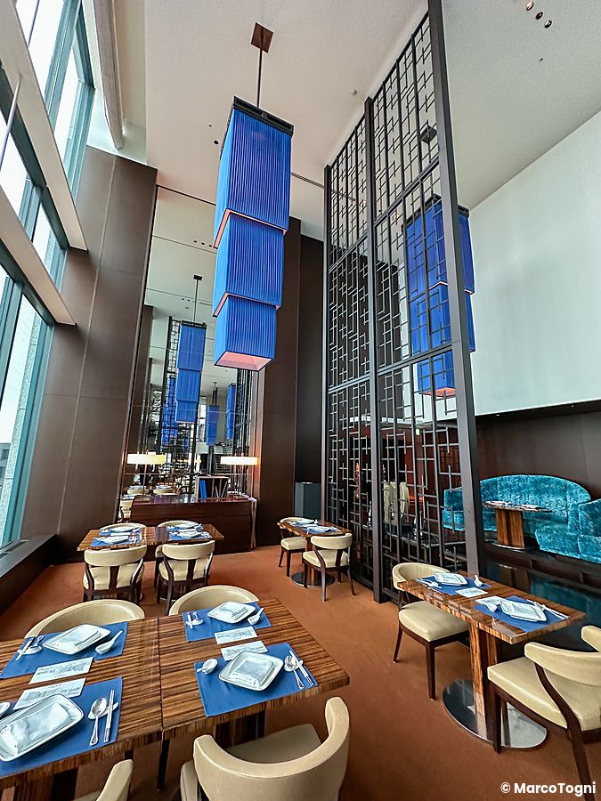 Tavoli apparecchiati all'interno della sala ristorante China Blue a Tokyo, caratterizzata da lampadari blu e ampie vetrate.