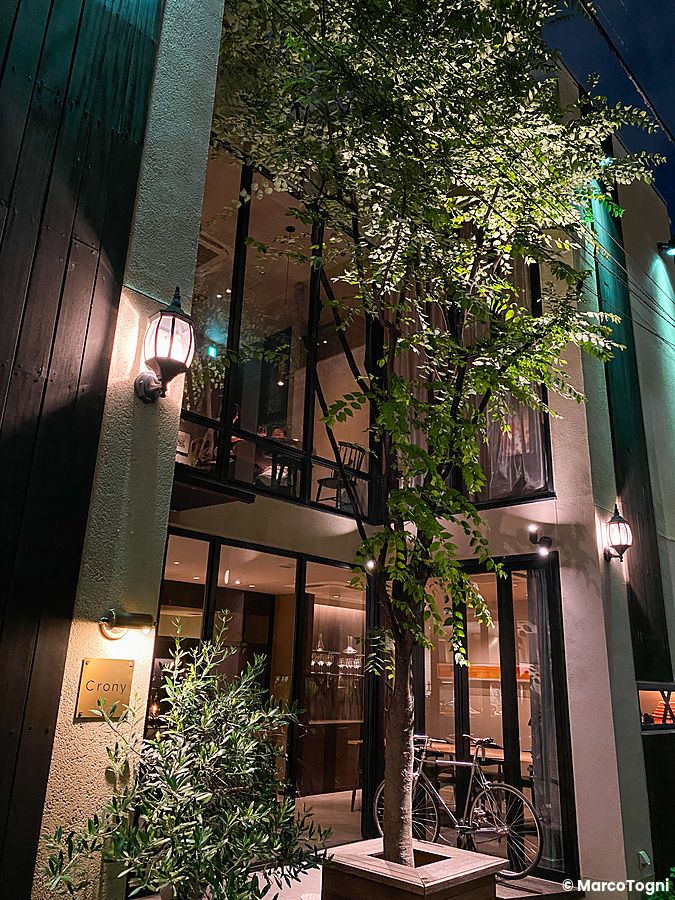L'esterno al Ristorante Crony a Tokyo di sera, con facciata moderna, ampie vetrate, un albero centrale e una bicicletta.