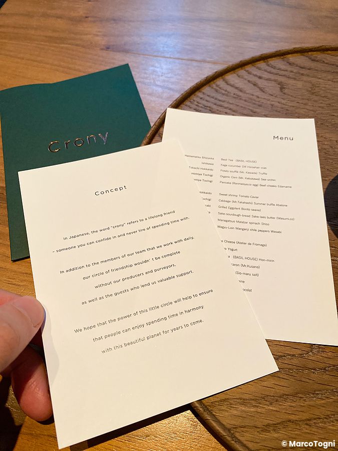 Fogli con il menu e la filosofia del Ristorante Crony, Tokyo, tenuti in mano sopra un tavolo in legno.