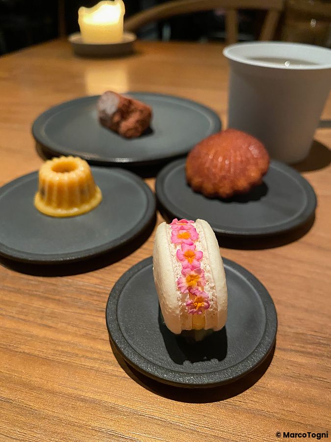 Piccola pasticceria servita su piattini neri al ristorante Crony, Tokyo, con un macaron floreale in primo piano.