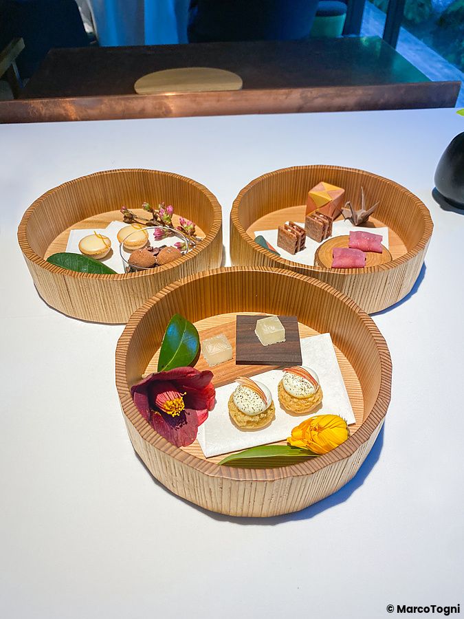 Tre contenitori in legno con piccola pasticceria a L'Effervescense, disposti su un tavolo bianco in un ristorante a Tokyo.