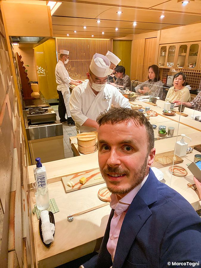 Selfie di Marco Togni seduto al bancone sushi a Sushi Ginza Onodera Souhonten con chef e clienti sullo sfondo.