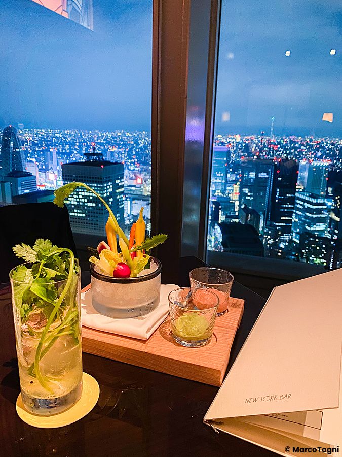 Cocktail e verdure su un tavolo con lo skyline notturno di Tokyo visibile da una grande vetrata del New York Bar.