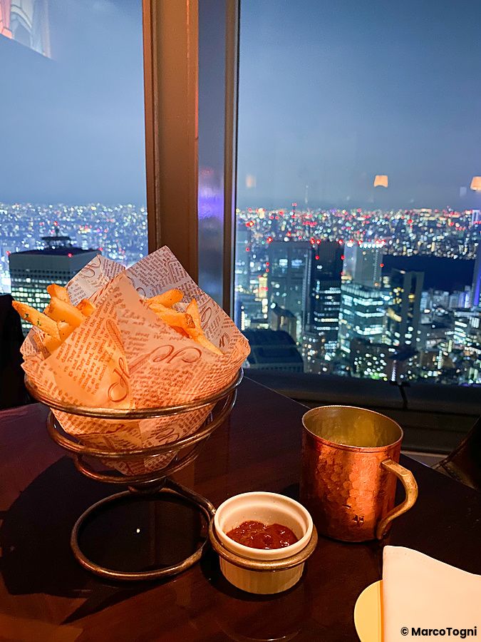 Patatine fritte in un cestino e una tazza in metallo su un tavolo al New York Bar, con la città di Tokyo sullo sfondo.