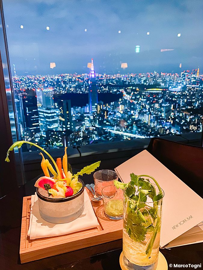 Veduta notturna di Tokyo dal New York Bar con un vassoio di verdure e un drink su un tavolo in primo piano.