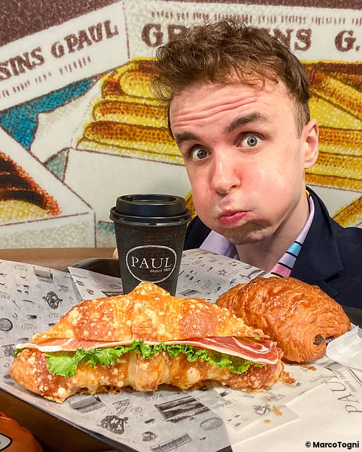 Marco Togni siede al tavolo di Paul davanti a un croissant salato, un dolce e un caffè da asporto.