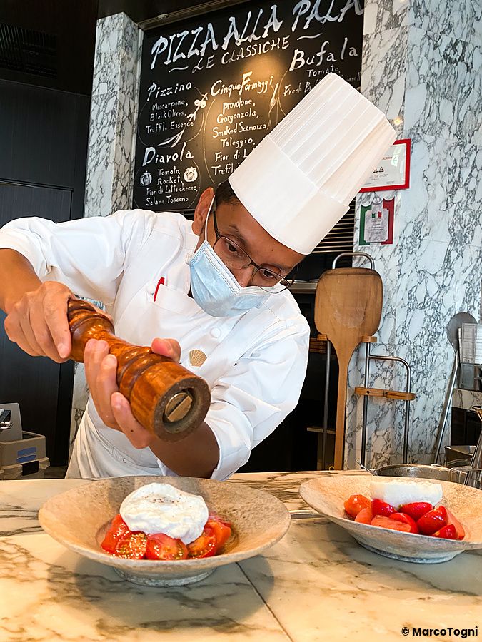 Un cuoco con divisa e mascherina macina pepe su una burrata con pomodoro a The Pizza Bar On 38th.