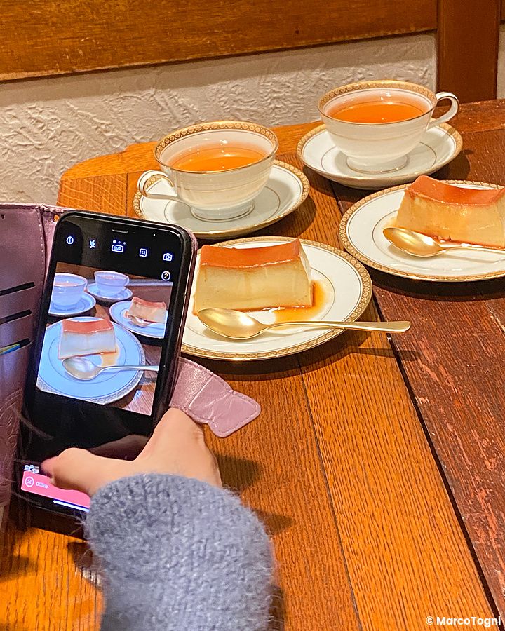 Una mano con un maglione grigio fotografa con uno smartphone budini e tazze di tè su un tavolo di legno nel caffè Satella.