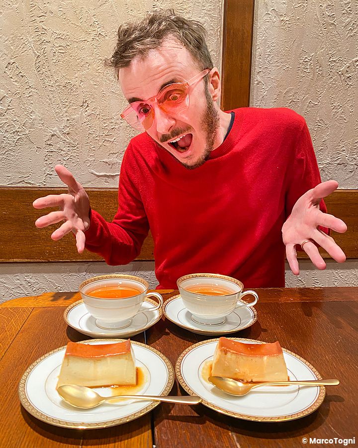 Marco Togni con maglione rosso e occhiali colorati siede a un tavolo al Satella a Shibuya davanti a budini e tazze.