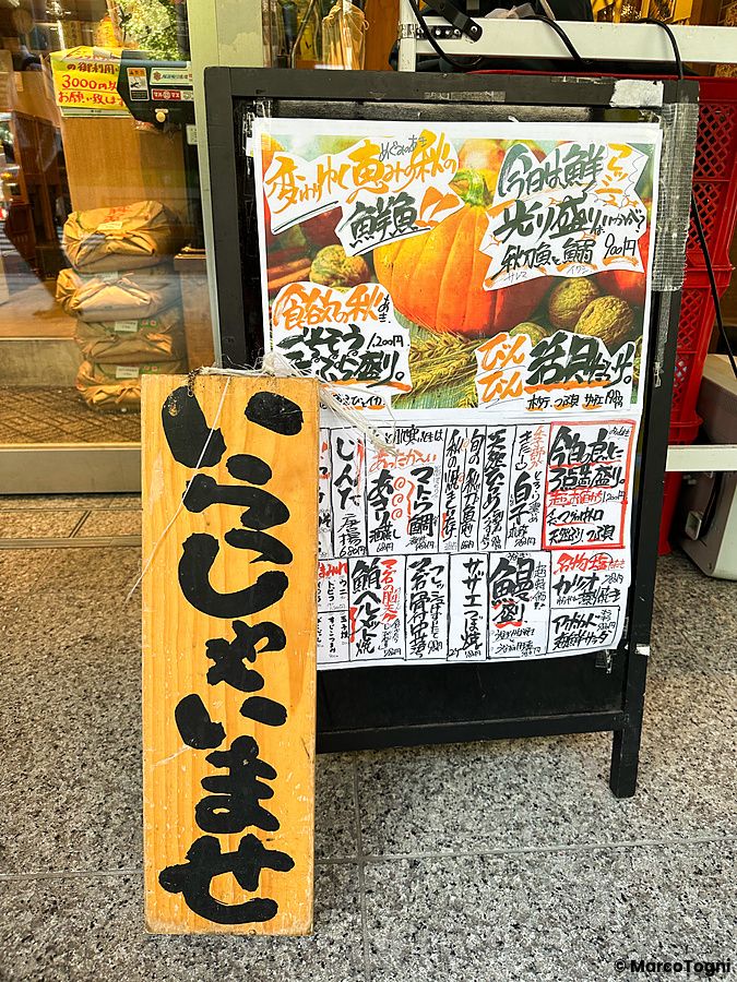 Cartello informativo con menu scritto a mano e targa in legno all'esterno del ristorante Sushi no Darihan a Shinjuku.