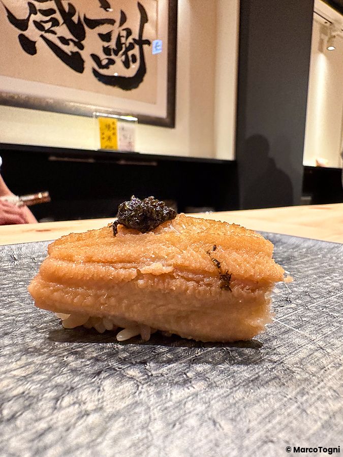 Un nigiri di anago con guarnizione scura posato su un piatto di pietra scura da Sushi Punch.