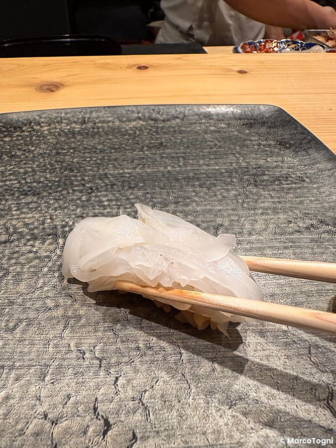 Un nigiri di aori ika sollevato con bacchette di legno sopra un piatto scuro ruvido da Sushi Punch a Tokyo.