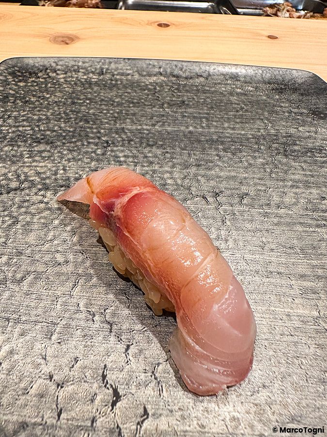 Un nigiri di isaki servito su un piatto scuro dalla texture irregolare presso Sushi Punch a Tokyo.