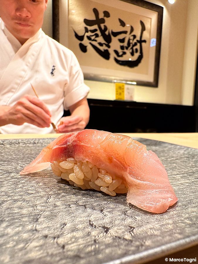 Un nigiri di isaki servito su un piatto scuro da Sushi Punch a Tokyo con un uomo in divisa da chef sullo sfondo.