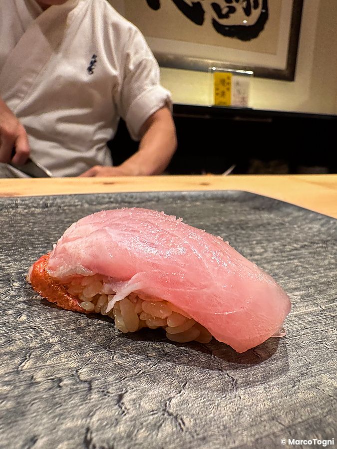 Un nigiri di kinmedai poggiato su un piatto di pietra scura con uno chef sullo sfondo da Sushi Punch.