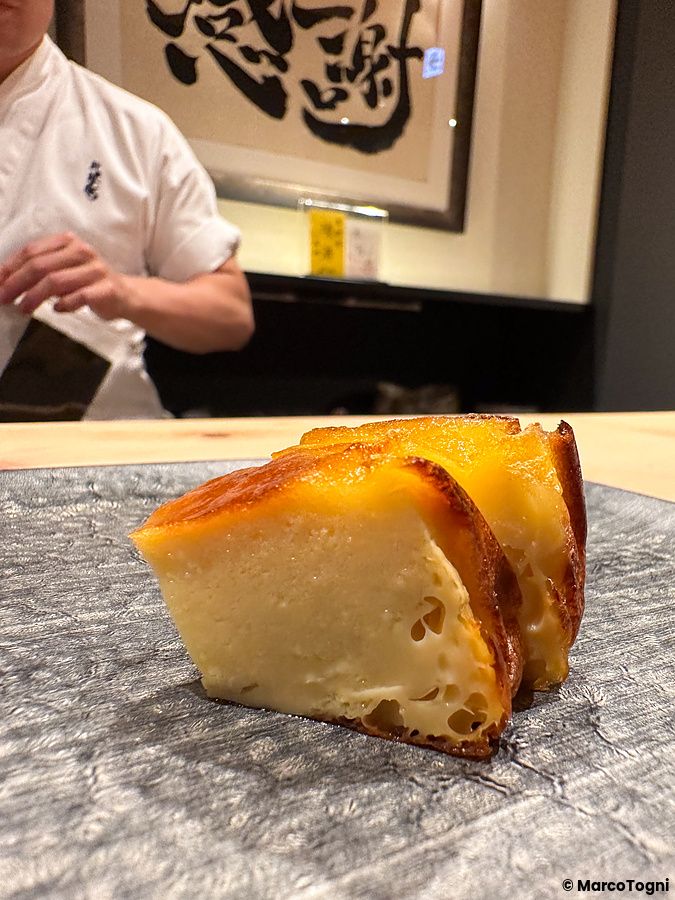 Un tamagoyaki dorato poggiato su un piatto scuro con uno chef in divisa bianca sullo sfondo da Sushi Punch.