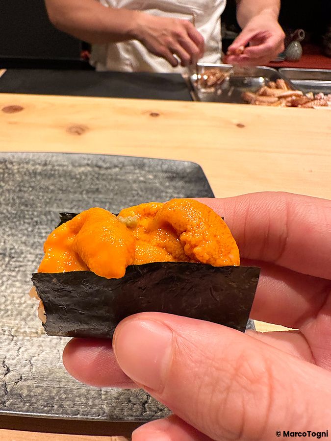 Una mano tiene un temaki con riccio preparato da Sushi Punch, con un cuoco che lavora sullo sfondo.