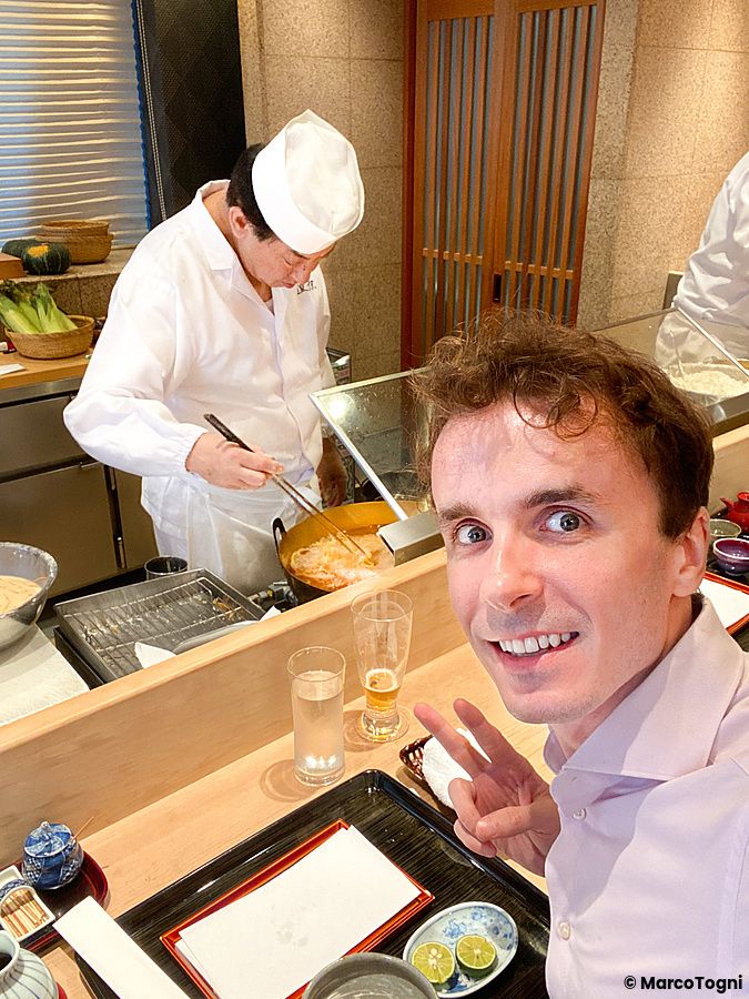 Marco Togni scatta un selfie mentre uno chef all'opera a Tempura Kondo cucina la tempura dietro il bancone in legno.