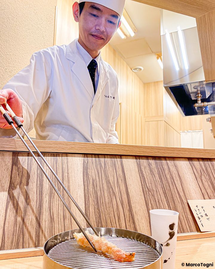 Un cuoco posiziona un gambero a Tempura Shimomura su una griglia metallica circolare sopra il bancone in legno.