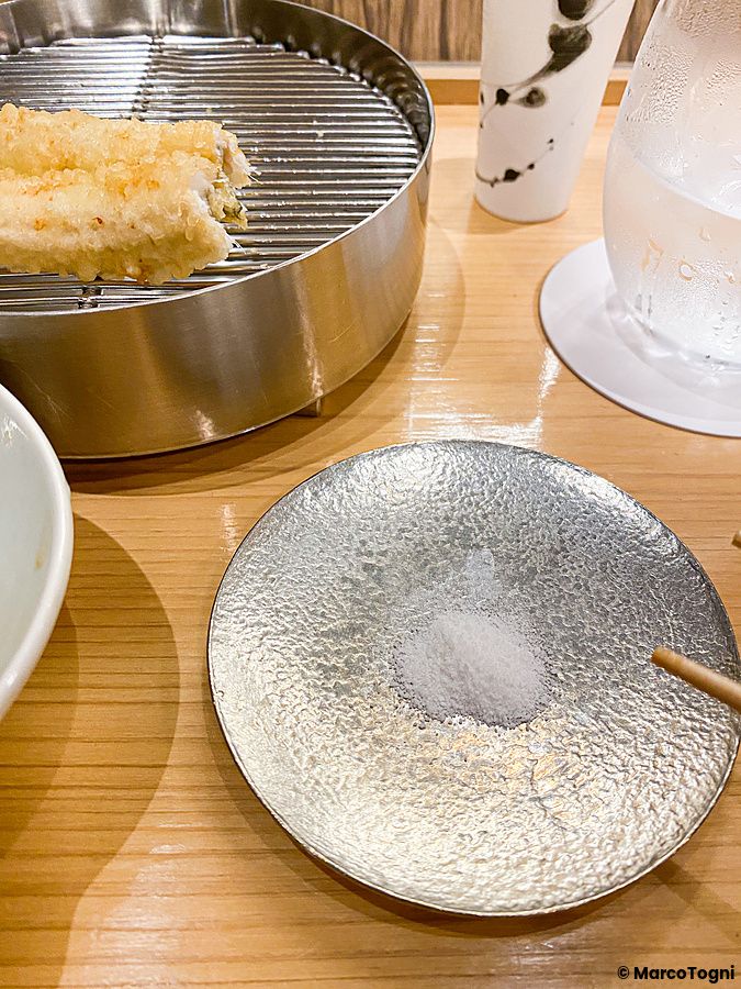 Un pezzo di tempura su una griglia e sale in un piattino metallico su un tavolo in legno al Tempura Shimomura a Tokyo.