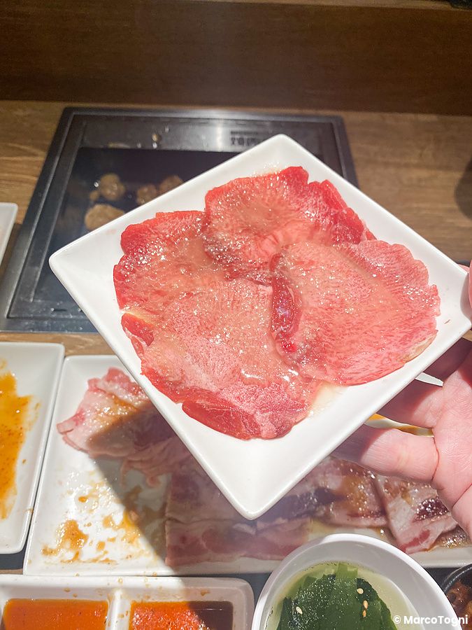 Una mano tiene un piattino con fettine di lingua di manzo cruda in un ristorante Yakiniku Like.