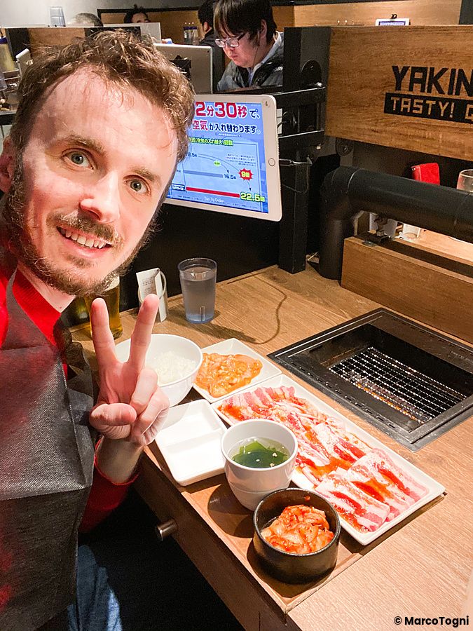 Marco Togni fa il segno di vittoria davanti a un tavolo piccolo con griglia a Yakiniku Like in un ristorante in Giappone.