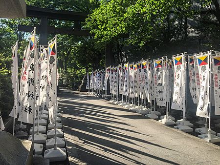 Una fila di bandiere bianche con scritte nere corre lungo il viale ombreggiato d'ingresso del Santuario Togo a Tokyo.
