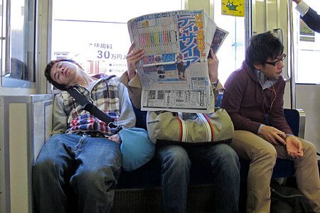 Persone sedute in un treno in Giappone, uno dorme, un altro legge un giornale.