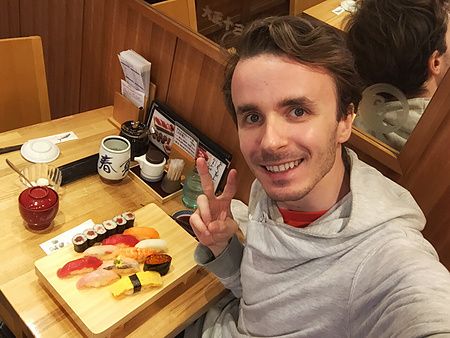 Marco Togni sorride facendo il gesto della vittoria davanti a un vassoio di sushi al Tama Sushi a Tachikawa.