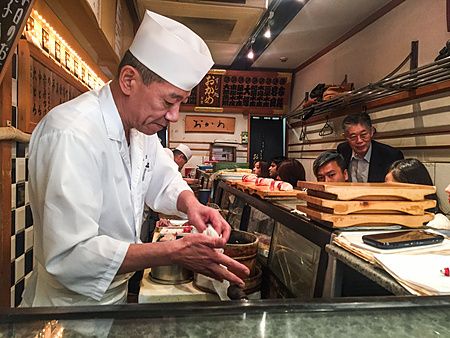 Uno chef prepara sushi nel Sushidokoro Okame, un ristorante nel mercato esterno di Tsukiji a Tokyo, con persone al bancone.