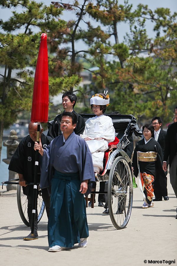 Una sposa in abito bianco tradizionale viaggia su un risciò trainato a Hiroshima, circondata da persone in abiti formali.