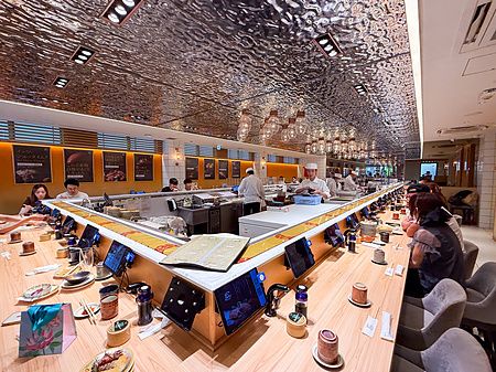 Interno moderno del Kaiten Sushi Ginza Onodera a Omotesando, con bancone a U, chef al lavoro e persone sedute ai tavoli.