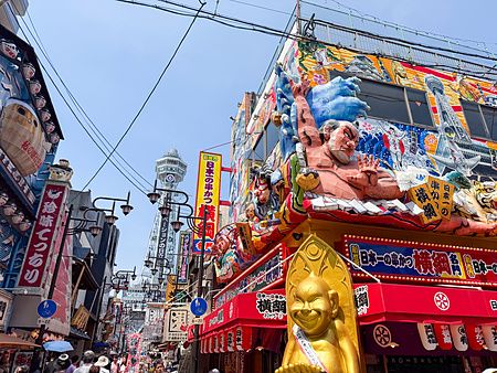 La torre Tsutenkaku si staglia sopra una via affollata nel quartiere di Shinsekai a Osaka, tra insegne e statue colorate.