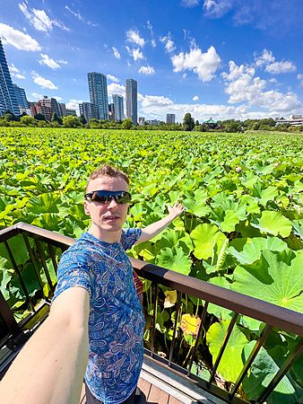 Marco Togni su passerella al Stagno Shinobazu di Ueno con foglie di loto e grattacieli.