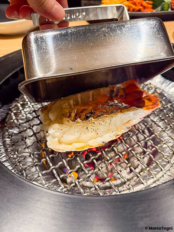 Una mano solleva un coperchio metallico su una griglia con braci ardenti che cuoce un crostaceo al Ristorante Toraji.