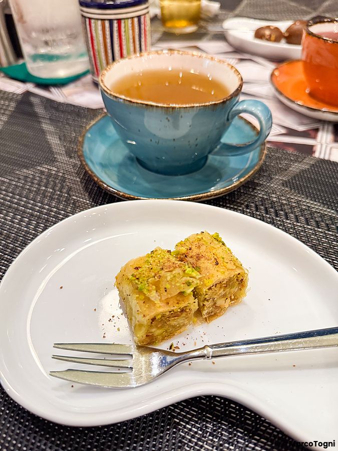 Una porzione di baklava a Seven Hills servita su un piatto bianco accanto a una tazza di tè azzurra su un tavolo.