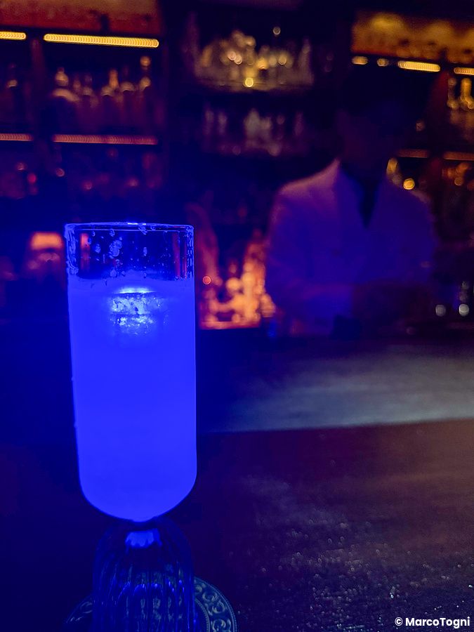 Un cocktail blu illuminato su un bancone del Bar Centifolia a Tokyo con una persona sfocata sullo sfondo.