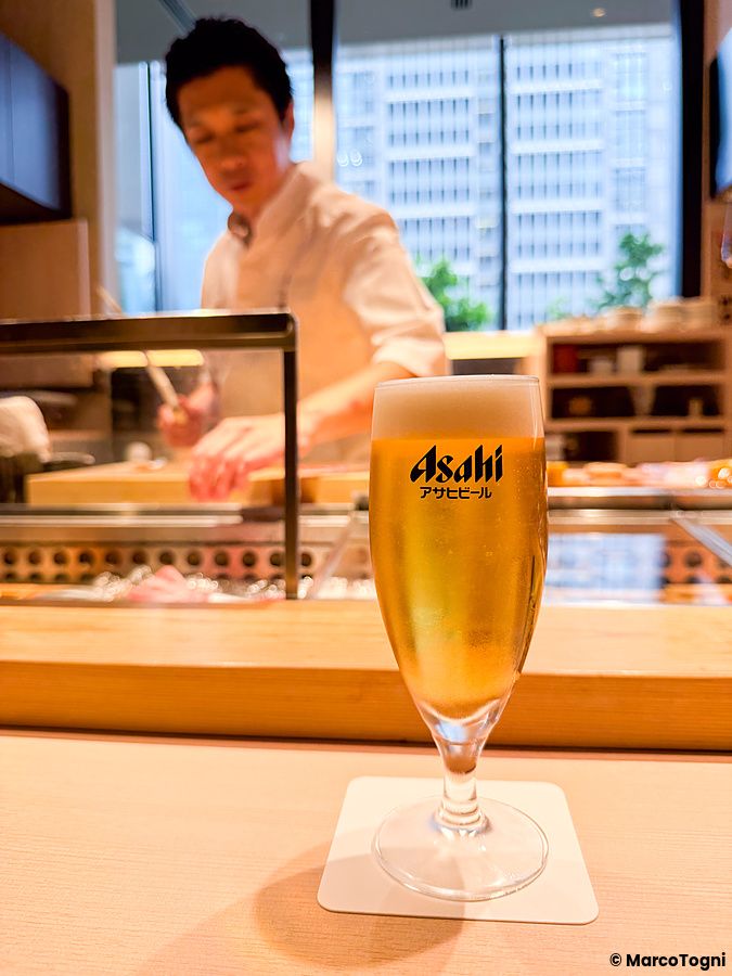 Un bicchiere di birra Asahi sul bancone del Sushi & Bar Fujiro Marunouchi Tokyo con uno chef sullo sfondo.