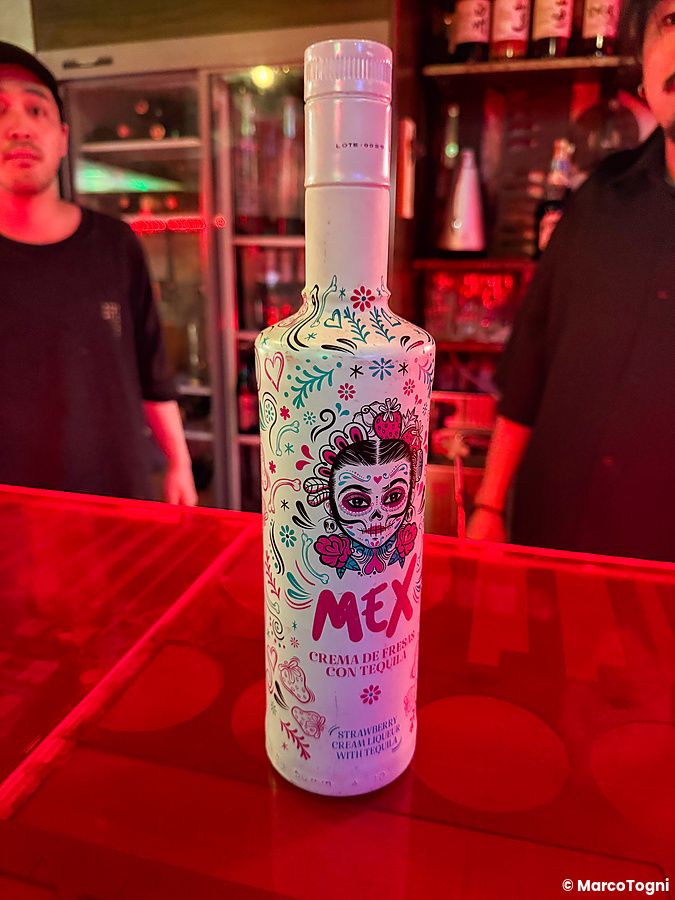 Bottiglia di liquore decorata su un bancone rosso al Neo Shinjuku Atsushi con due uomini sullo sfondo.
