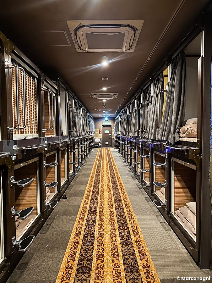 Corridoio interno del capsule hotel Anshin Oyado a Shinjuku con due file di capsule letto e moquette decorata.