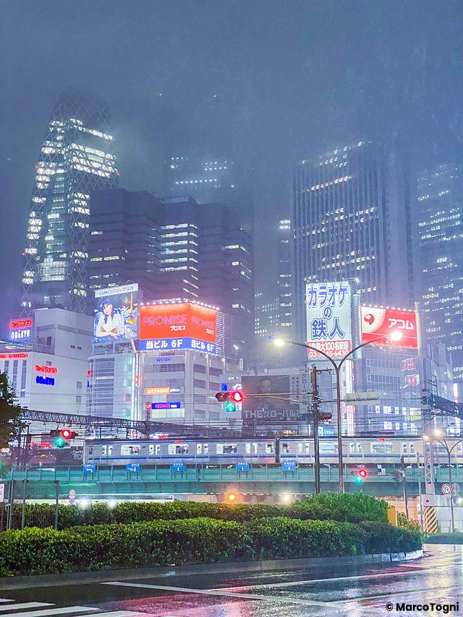 Veduta notturna dei grattacieli di Shinjuku a Tokyo, con un treno in transito su binari sopraelevati sotto la pioggia.