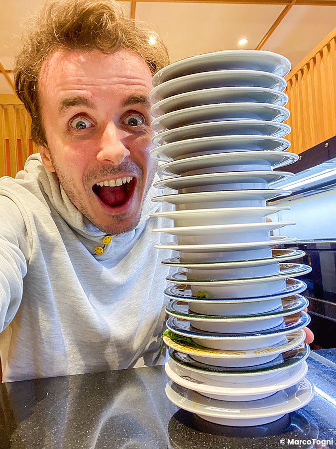 Marco Togni sorride accanto a una torre di piatti vuoti in un kaiten sushi in Giappone.
