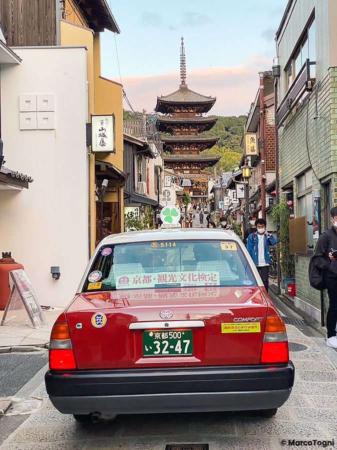 Un taxi rosso con targa giapponese fermo su una strada nel quartiere Higashiyama con la pagoda Yasaka sullo sfondo.