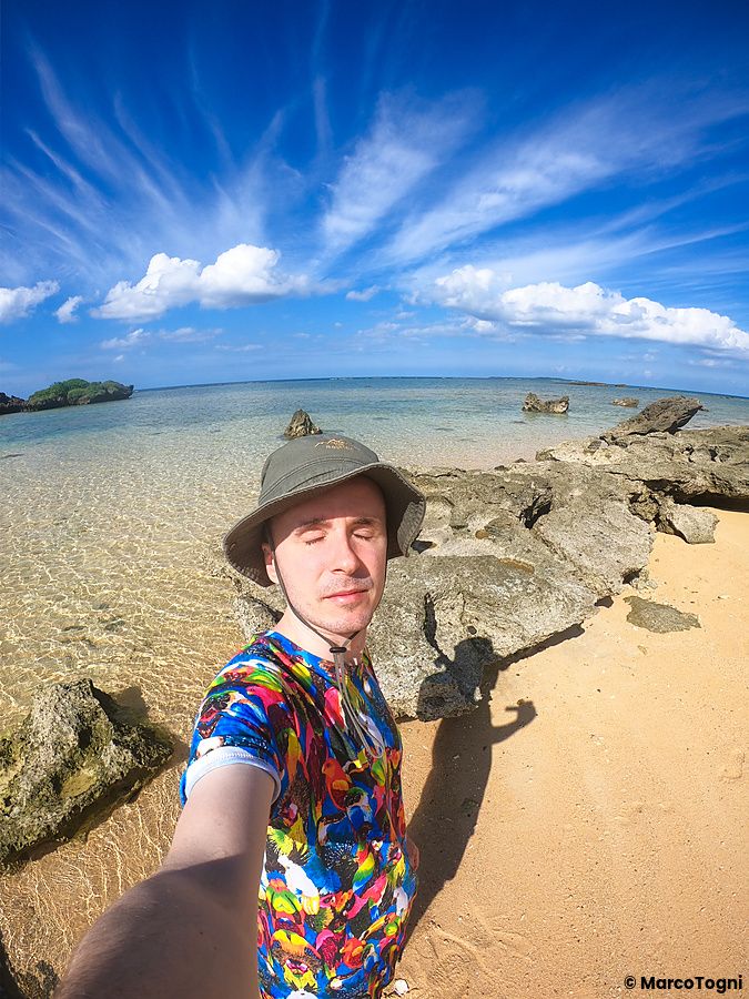 Marco Togni con cappello e maglietta colorata scatta un selfie sulla sabbia di Hoshizuna Beach, a Okinawa.