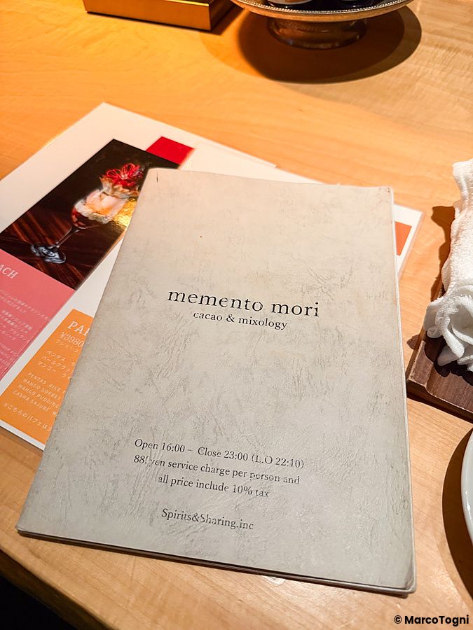 Un menù cartaceo con la scritta Memento Mori cacao & mixology posto su un tavolo in legno all'interno del locale.