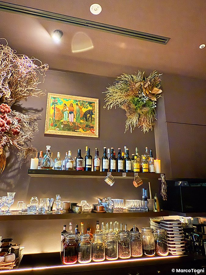 Ripiano di un bar a Tokyo con vasi di chicchi di cacao, bottiglie di liquore, fiori secchi e un quadro sulla parete.