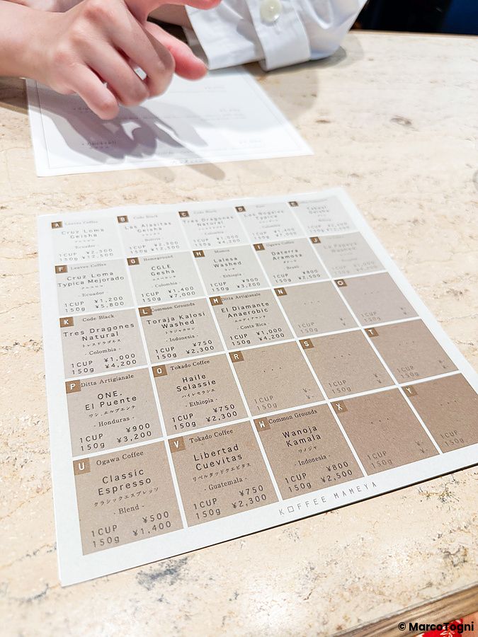 Il menu coi vari tipi di caffè a Koffee Mameya Kakeru appoggiato su un bancone, con una persona visibile sullo sfondo.
