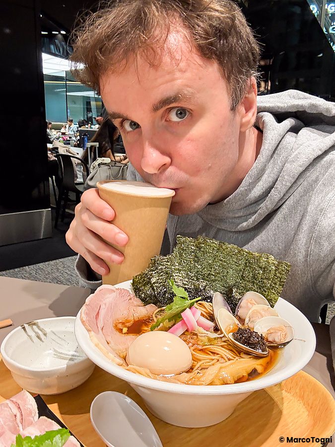 Marco Togni beve da un bicchiere di carta davanti a una ciotola di ramen con vongole presso il ristorante Mugi to Olive.