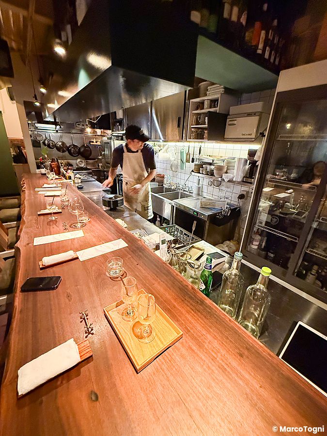 Interno a Mushroom Tokyo con un lungo bancone in legno e un individuo al lavoro nella cucina in acciaio.